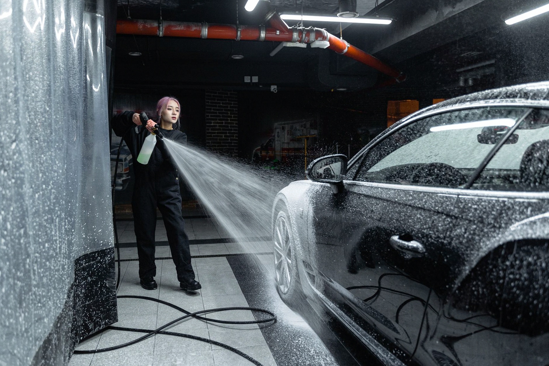 Woman using a pressure washer to clean a black car in an indoor car wash setting.